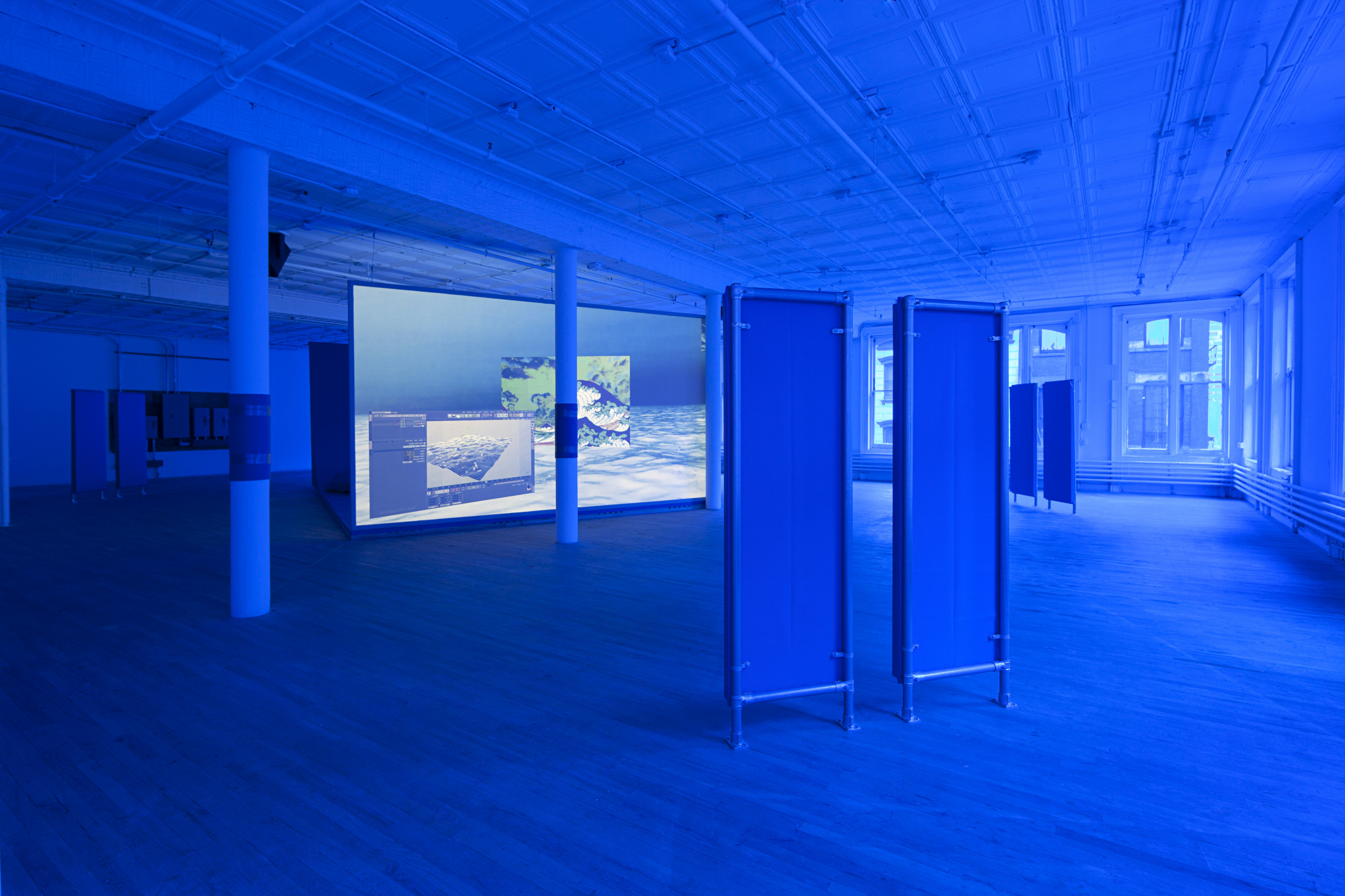 An angled view of a large video monitor screen positioned on the ground behind a collumn. The room is tinted blue. Three pairs of pipe-frame panels visible in the front and both sides of the screen, which projects a video-still of a composited frame that includes computer generated images of the ocean.