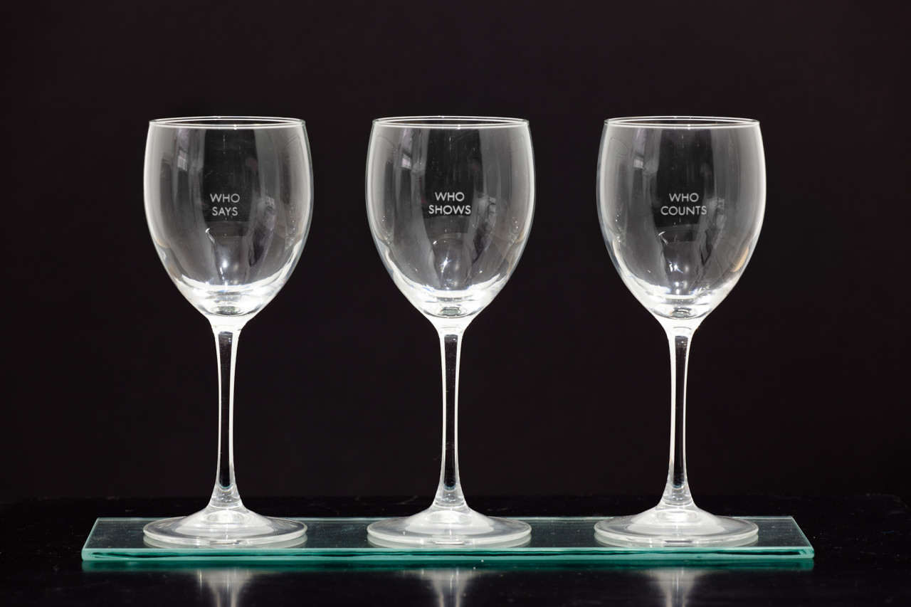 Three empty wine glasses in a row, each engraved with two words: "who says," "who shows," "who counts."