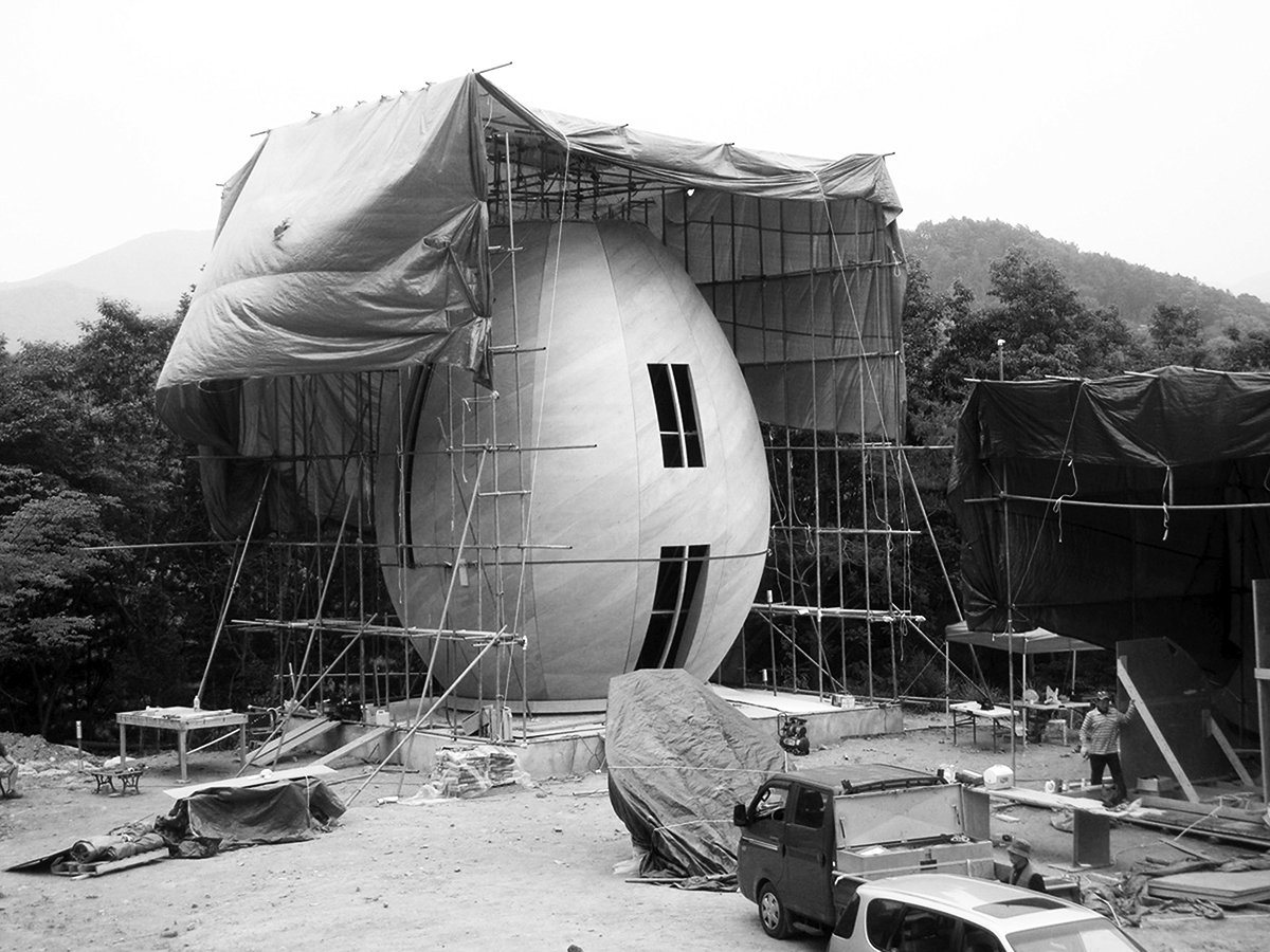 A black and white photograph showing the construction of a egg-shaped structure. Scaffolding is set up around it, and a window and door are cut out from the front