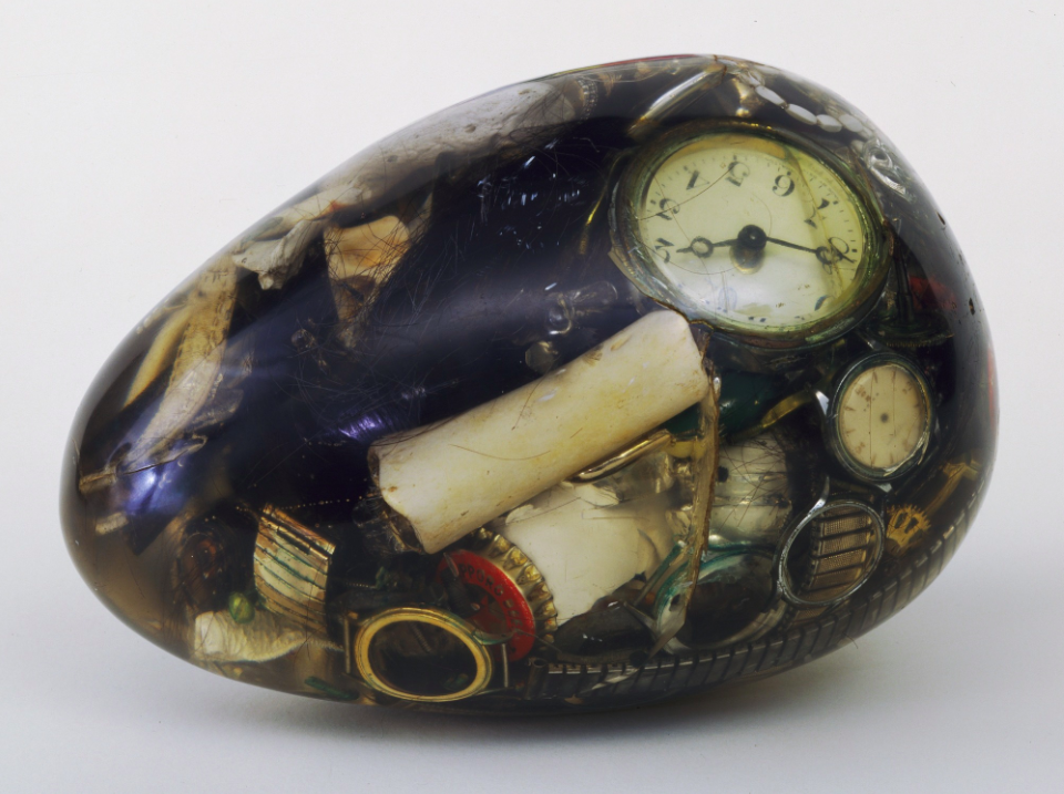 A transparent egg filled with junk: old watch faces, a cigarette butt, bottle caps, crumpled papers