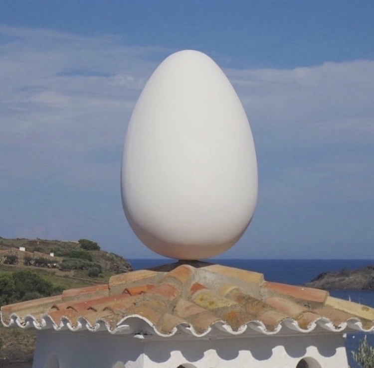 A circular terracotta roof with an enormous white egg sitting atop it, as if balancing in place. Beyond the roof is the ocean and a bright blue sky