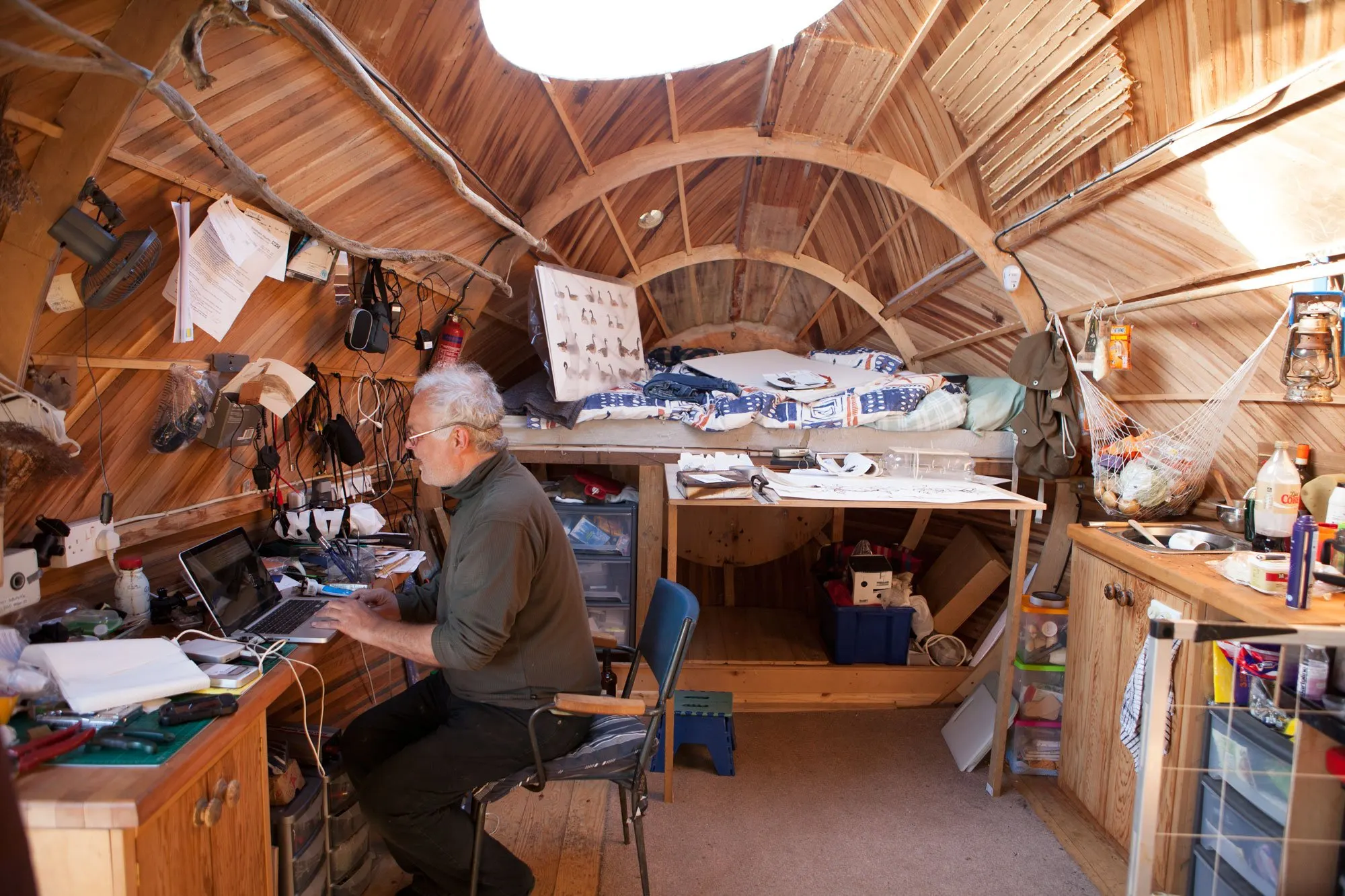 A man sits inside a homey oval wooden structure that almost looks like the hull of a ship. He works from a laptop on a desk strewn with stuff. Behind him is a kitchenette and beyond him looks like a living area