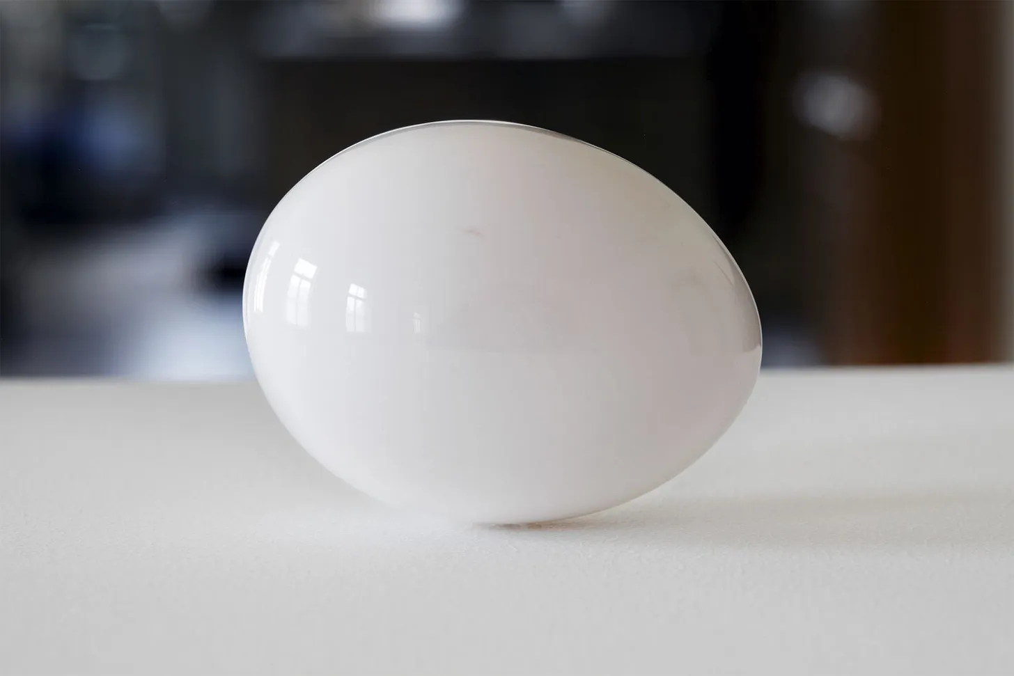 A white egg without a shell sits on a white table. The egg is so shiny you can see the windows of the surrounding room in its reflection