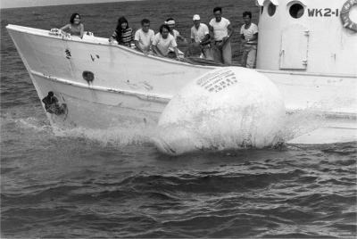 A black and white photograph of the boat passing the floating egg structure. The people on the boat look at it from the deck