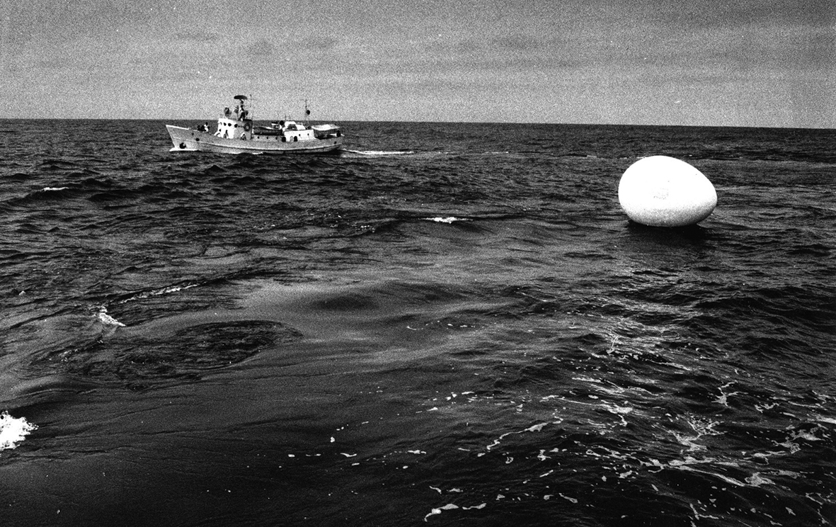 A black and white photograph of the ocean, a boat in the background, and the egg sculpture floating on the water in the foreground