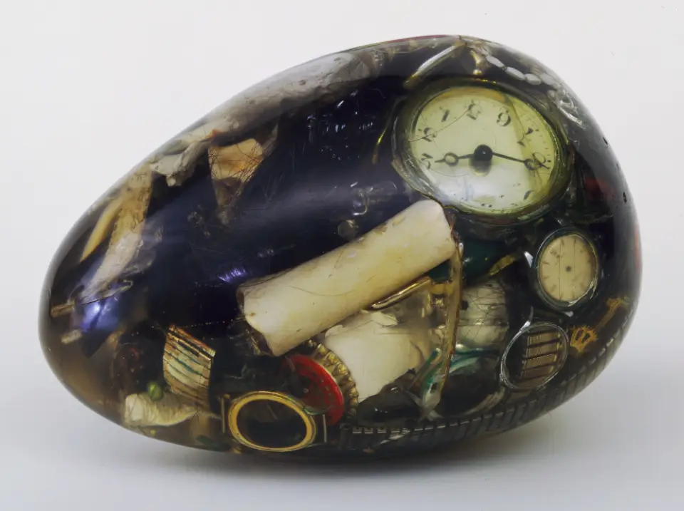 A transparent egg filled with junk: old watch faces, a cigarette butt, bottle caps, crumpled papers