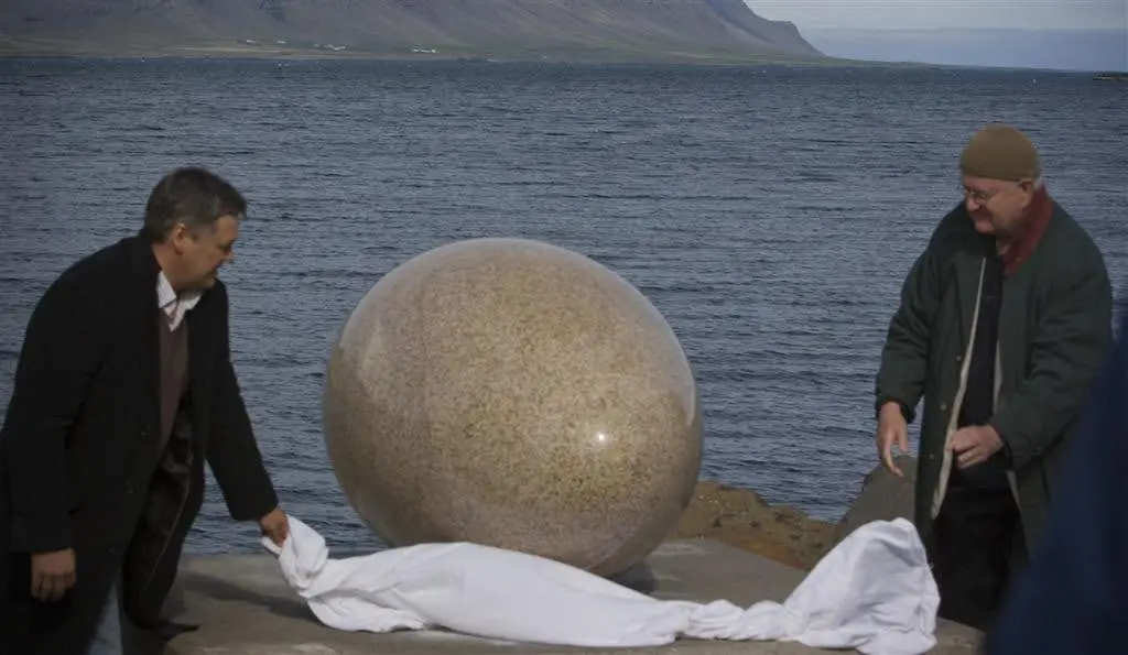Two men pull a white sheet off a large granite egg, as if revealing it for the first time. The ocean can be seen in the background